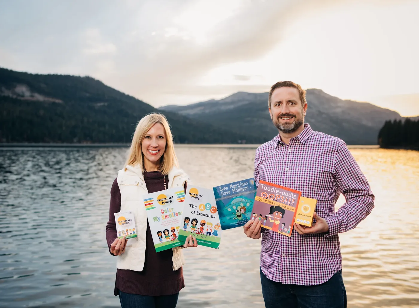 Bryan Jones and Cami Childs standing by Donner Lake holding the games and books they have made, including Feelings and Dealings, Color My Emotions, The ABCs of Emotions, Even Martians Have Manners and Toodle-oodle Oops